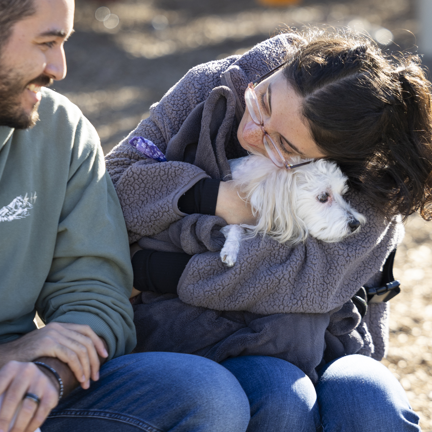 a woman hugs a small white dog sitting in her lap while a man sitting next to her smiles at them