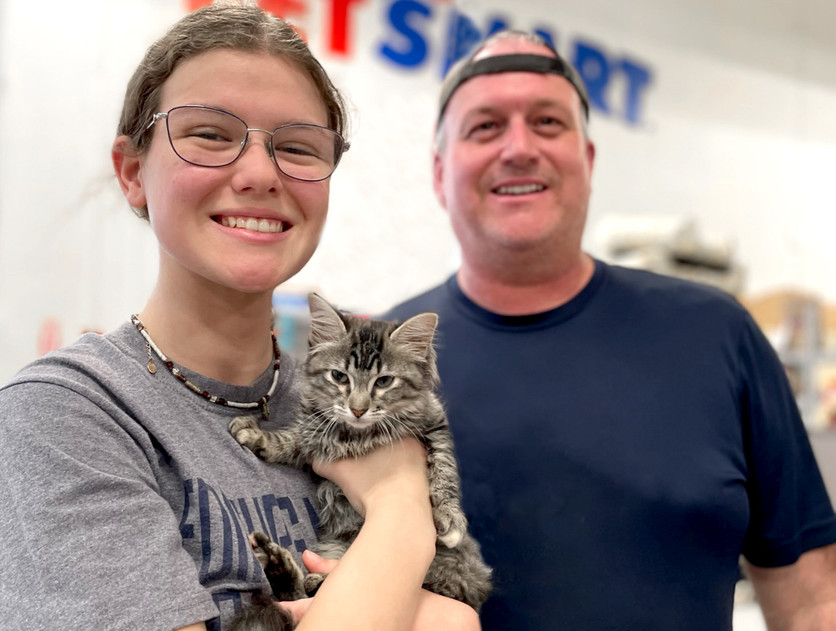 young woman smiling and wearing glasses holds a kitten in her arms. She stands next to a man wearing a backwards baseball cap.