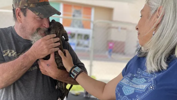 a man wearing a baseball cap holds a puppy in his arms while a woman reaches over to pet the puppy